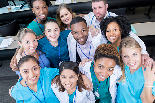 This is a view from above of a group of medical students looking up and smiling for the camera.  They are standing in a lecture hall with table behind them.